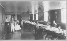 SA0145 - The Shaker women, also including Sarah Burger, were of the North Family. Photo is of  long tables set for a meal and a tree or bush branches hanging from the ceiling. An ad on the back is for other views., Winterthur Shaker Photograph and Post Card Collection 1851 to 1921c
